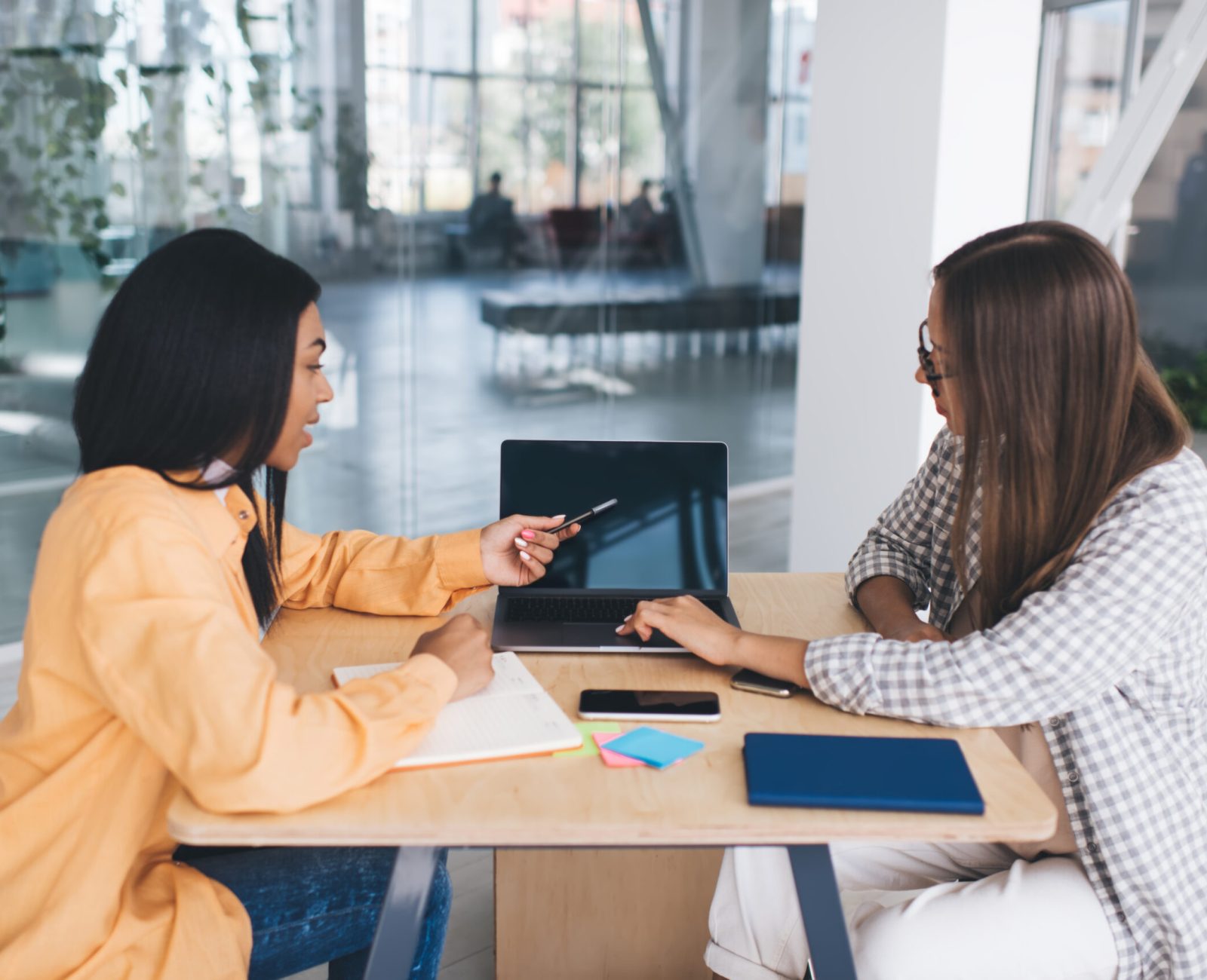 Black businesswoman showing something on laptop to her european female colleague. Concept of modern successful women. Idea of teamwork. Young girls sitting at wooden desk in office